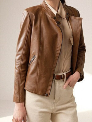 Woman standing in a casual pose wearing a brown leather jacket over a beige shirt and cream pants, holding a small clutch handbag.